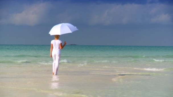 Serene jonge vrouw met een witte parasol in het ondiepe water naast een prachtige witte zandstrand — Stockvideo