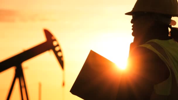 Silhouette einer Ingenieurin mit Klemmbrett, die mit einem Handy den Ort der Erdölförderung bei Sonnenuntergang überwacht — Stockvideo