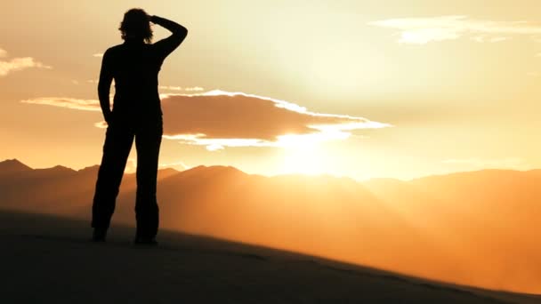 Одинокая женщина в силуэте путешествует по песчаным дюнам и наблюдает заход солнца — стоковое видео