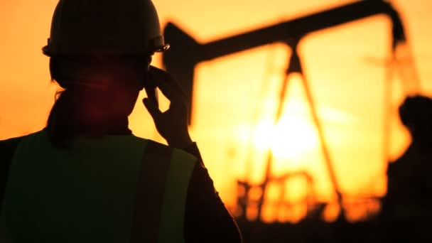 Silhouet van vrouwelijke ingenieur met Klembord met behulp van een mobiele telefoon toezicht op de site van ruwe olieproductie bij zonsondergang — Stockvideo