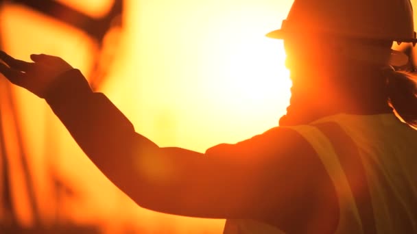 Silhueta de engenheira com prancheta usando um telefone celular supervisionando o local de produção de petróleo bruto ao pôr do sol — Vídeo de Stock