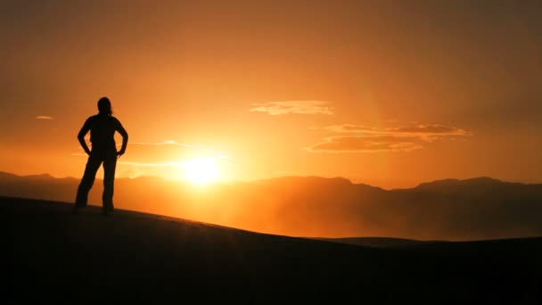 Solitary female in silhouette hiking across sand dunes & watching the sun setting — Stock Video