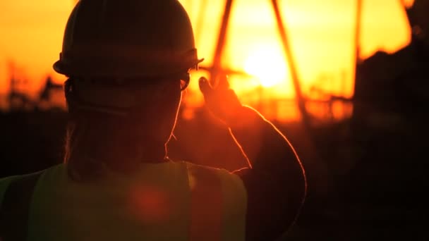 Silhueta de engenheira com prancheta usando um telefone celular supervisionando o local de produção de petróleo bruto ao pôr do sol — Vídeo de Stock