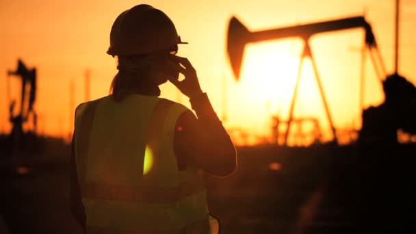 Силуэт женщины-инженера с буфером обмена с помощью мобильного телефона, контролирующего место добычи нефти на закате — стоковое видео