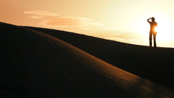 Solitary female in silhouette hiking across sand dunes & watching the sun setting — Stock Video