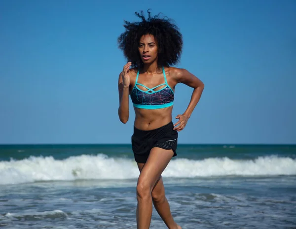 スポーツウェアを着たアフリカ系アメリカ人女性が海の波を越えて心臓の健康のためにビーチで楽しいワークアウトを楽しむ — ストック写真