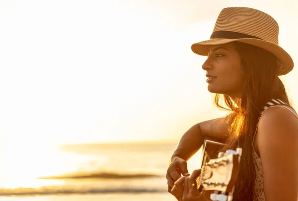 Smiling young Indian woman wearing hat on relaxing Summer vacation playing guitar music on beach at sunrise