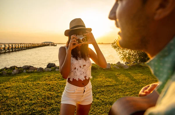 若い民族の女性は帽子をかぶって休暇でパートナーの写真を撮るギターで海で日没時に演奏 — ストック写真