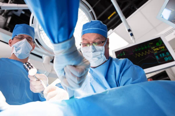 病院の劇場で手術を行う専門医療チームと男性白人外科医の背後にある患者の重要な兆候を示すモニター — ストック写真