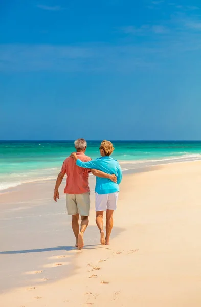 Loving seniors walking barefoot on sandy beach with arms embraced enjoying freedom in retirement on tropical island Bahamas