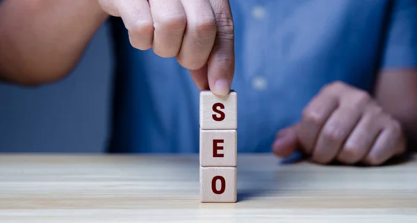 Businessman's hand picks up a wooden block with wooden letters for SEO on a wooden table (Search Engine Optimization).