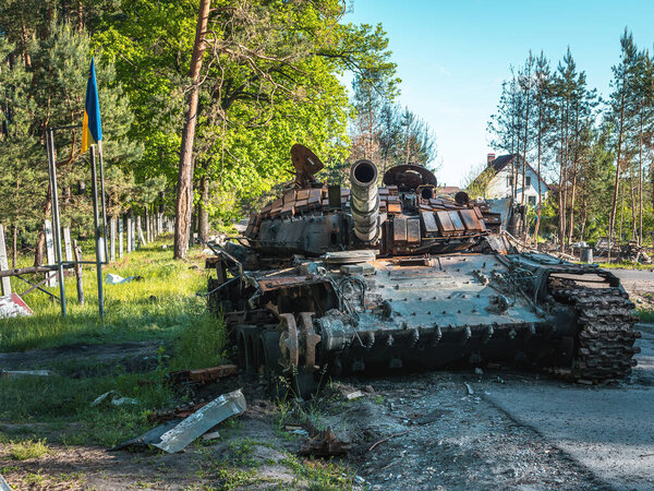 War in Ukraine, a destroyed Russian tank stands next to the Ukrainian flag on the background of the forest, front view.