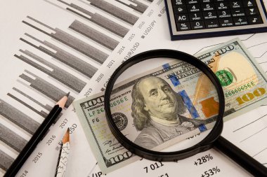 Bir büyüteç altındaki 100 dolarlık banknot, hisse senedi tabloları ve alıntılarına karşı. İş ve Finans