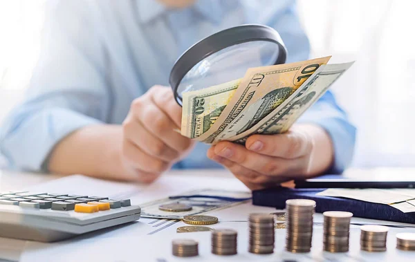 Bir adam büyüteçle parayı inceler. Büyüteç altında dolar. Mali işler. Gelir ve kar