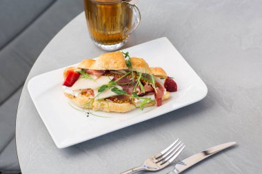 Prosciutto, böğürtlen, dijon hardalı, peynir ve beyaz tabakta bir fincan çay ile kruvasan sandviçi. Avrupai kahvaltı.