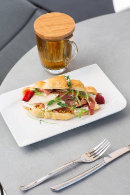 Prosciutto, böğürtlen, dijon hardalı, peynir ve beyaz tabakta bir fincan çay ile kruvasan sandviçi. Avrupai kahvaltı.