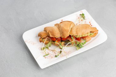 Avokadolu kruvasan sandviçi, beyaz tabakta domatesler ve parlak bir arka plan. Avrupai kahvaltı.