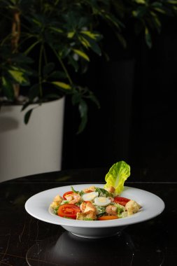 Izgara karides, domates, haşlanmış yumurta, marul, kızarmış ekmek ve yeşil sos ile Sezar salatası. Kahverengi yuvarlak bir tabağın üst görüntüsü. Taze sebzeler ve karanlık bir arka planda çatal bıçak takımı..