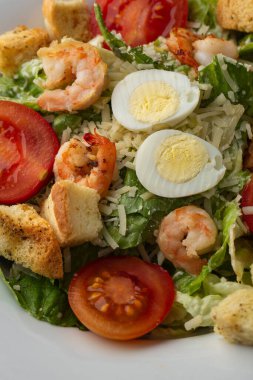 Izgara karides, domates, haşlanmış yumurta, marul, kızarmış ekmek ve yeşil sos ile Sezar salatası. Kahverengi yuvarlak bir tabağın üst görüntüsü. Taze sebzeler ve karanlık bir arka planda çatal bıçak takımı..