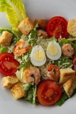 Izgara karides, domates, haşlanmış yumurta, marul, kızarmış ekmek ve yeşil sos ile Sezar salatası. Kahverengi yuvarlak bir tabağın üst görüntüsü. Taze sebzeler ve karanlık bir arka planda çatal bıçak takımı..