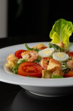 Izgara karides, domates, haşlanmış yumurta, marul, kızarmış ekmek ve yeşil sos ile Sezar salatası. Kahverengi yuvarlak bir tabağın üst görüntüsü. Taze sebzeler ve karanlık bir arka planda çatal bıçak takımı..