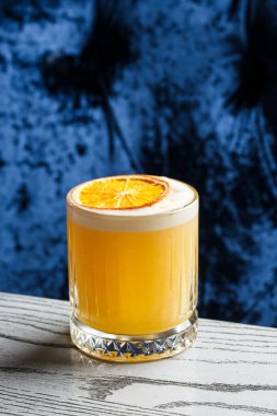 Beyaz köpüklü sarı bir kokteyl. Büyük bir buz küpü. Mavi bulanık arka planda turuncu cipslerle süslenmiş..