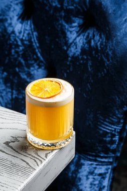 Beyaz köpüklü sarı bir kokteyl. Büyük bir buz küpü. Mavi bulanık arka planda turuncu cipslerle süslenmiş..