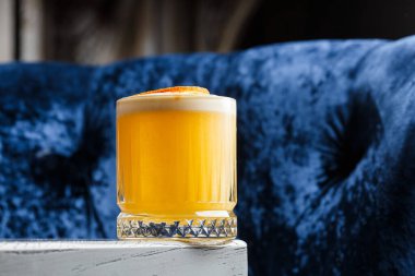 Beyaz köpüklü sarı bir kokteyl. Büyük bir buz küpü. Mavi bulanık arka planda turuncu cipslerle süslenmiş..