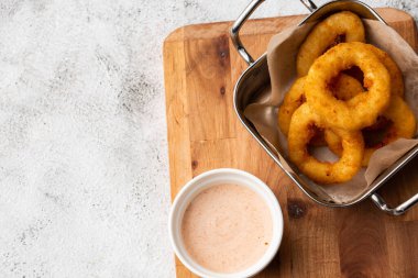 Derin kızartılmış soğan halkaları metal sepetin içinde, ahşap tahtada beyaz sosla servis ediliyor mermer arka planda.