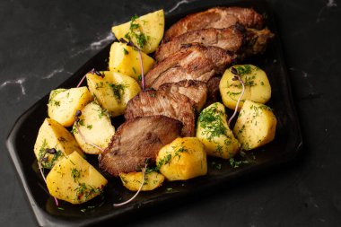 Fırında taze patates, deniz tuzu ve domuz filetosu ile mermer masada siyah bir tabakta parçalara ayrılmış.