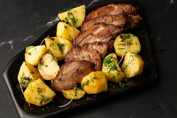Fırında taze patates, deniz tuzu ve domuz filetosu ile mermer masada siyah bir tabakta parçalara ayrılmış.