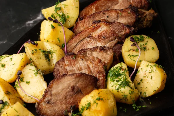 Fırında taze patates, deniz tuzu ve domuz filetosu ile mermer masada siyah bir tabakta parçalara ayrılmış.