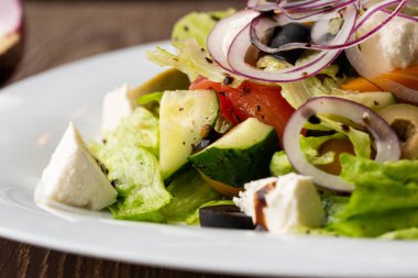 Taze sebzeli Yunan salatası: domates, salatalık, kırmızı biber, marul, soğan, zeytin ve peynir. Tahta bir arka planda beyaz yuvarlak bir plakaya yakın plan.