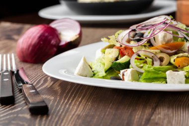 Taze sebzeli Yunan salatası: domates, salatalık, kırmızı biber, marul, soğan, zeytin ve peynir. Tahta bir arka planda beyaz yuvarlak bir plakaya yakın plan.
