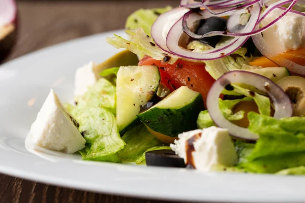 Taze sebzeli Yunan salatası: domates, salatalık, kırmızı biber, marul, soğan, zeytin ve peynir. Tahta bir arka planda beyaz yuvarlak bir plakaya yakın plan.