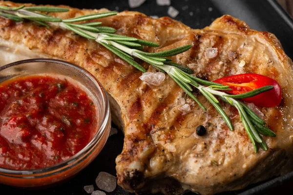 Roasted pork chop steak with spices, rosemary and red sauce on black plate on wooden background. Pork, beef, chop on a bone. top view. place for text