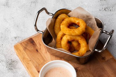 Derin kızartılmış soğan halkaları metal sepetin içinde, ahşap tahtada beyaz sosla servis ediliyor mermer arka planda.