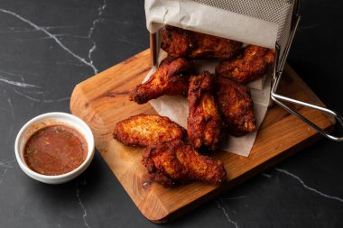 Gerçek sıcak barbekü sosuyla kaplanmış kızarmış tavuk kanatları mermer arka planda kesme tahtasında kırmızı acı biber sosuyla servis edilir.