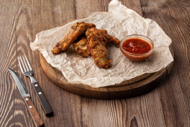 Hakiki sıcak barbekü sosuyla kaplanmış kızarmış tavuk kanatları. Ahşap arka planda parşömen kağıdında kırmızı acı biber sosuyla servis ediliyor.