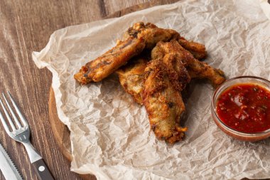 Hakiki sıcak barbekü sosuyla kaplanmış kızarmış tavuk kanatları. Ahşap arka planda parşömen kağıdında kırmızı acı biber sosuyla servis ediliyor.