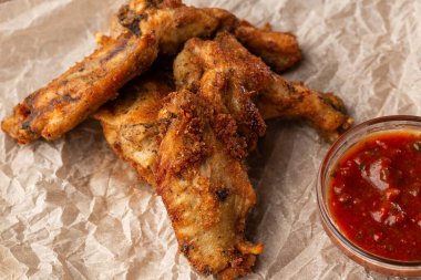 Hakiki sıcak barbekü sosuyla kaplanmış kızarmış tavuk kanatları. Ahşap arka planda parşömen kağıdında kırmızı acı biber sosuyla servis ediliyor.