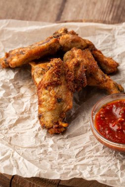Hakiki sıcak barbekü sosuyla kaplanmış kızarmış tavuk kanatları. Ahşap arka planda parşömen kağıdında kırmızı acı biber sosuyla servis ediliyor.