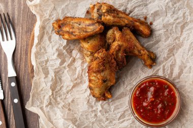 Hakiki sıcak barbekü sosuyla kaplanmış kızarmış tavuk kanatları. Ahşap arka planda parşömen kağıdında kırmızı acı biber sosuyla servis ediliyor.