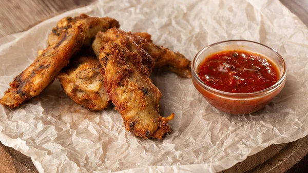 Hakiki sıcak barbekü sosuyla kaplanmış kızarmış tavuk kanatları. Ahşap arka planda parşömen kağıdında kırmızı acı biber sosuyla servis ediliyor.