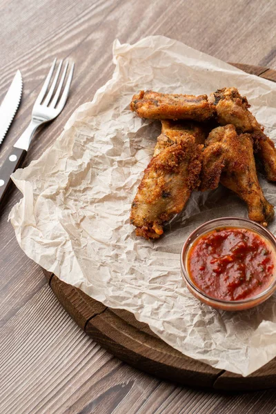 Hakiki sıcak barbekü sosuyla kaplanmış kızarmış tavuk kanatları. Ahşap arka planda parşömen kağıdında kırmızı acı biber sosuyla servis ediliyor.