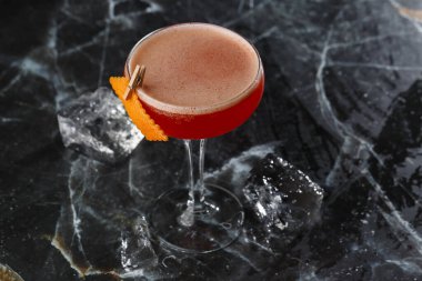 Portakal kabuğuyla süslenmiş bir coupe bardağında kırmızı ekşi kokteyl. Clover Club ve ithal cin. Siyah mermer arka planda.