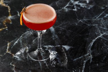 Portakal kabuğuyla süslenmiş bir coupe bardağında kırmızı ekşi kokteyl. Clover Club ve ithal cin. Siyah mermer arka planda.