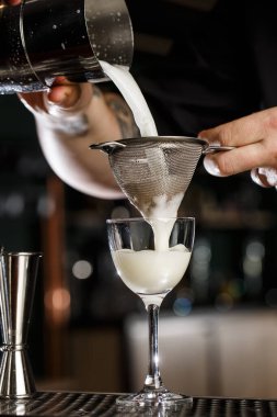 Adam barmen dikkatli bir şekilde Pisco ekşi kokteylini çelik karıştırıcıdan elek yoluyla cama filtreler. Yakın çekim görünümü