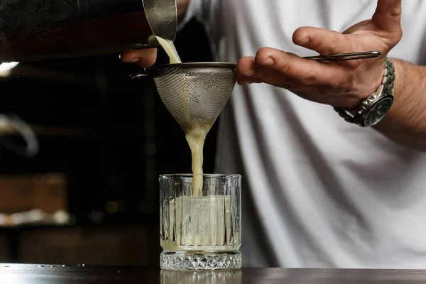 man bartender carefully filters bright alcoholic cocktail from steel shaker cup into glass through sieve. Close-up view