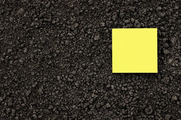 Yellow note paper square on black soil texture ground close up. Yellow sticky note blank paper flat soil organic earth texture. Mockup on ground soil background top view from above. Humus. Agriculture